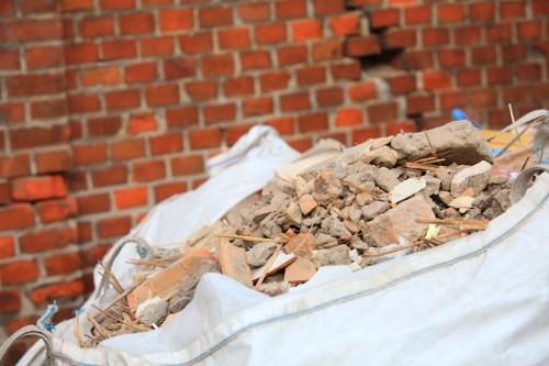 A large white construction or rubble bag filled with a mix of broken bricks, concrete chunks, and debris, placed outdoors on a paved surface in front of a red brick wall. The rubble appears to consist of irregularly shaped stone and brick fragments with rough textures and earthy tones, including shades of beige, brown, and reddish-orange. The bag is open at the top, revealing the heavy, coarse materials inside, and some small wooden or plastic tools are partially visible among the debris. The environment suggests a tidy garden clearance or building waste removal scene that could be associated with services provided by Garden Clearance Colindale, located near Colindale, North London, in the northwest postcode area. The scene emphasizes the handling and disposal of construction waste in a residential or garden setting, with natural daylight illuminating the materials clearly for visual accuracy related to rubbish removal services in the area.