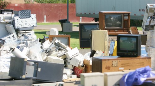 A large outdoor pile of discarded electronic waste and obsolete appliances, including multiple old CRT televisions, computer monitors, and various boxed and unboxed devices. The electronics are stacked haphazardly on a paved surface, with some screens facing outward and others tilted or partially obscured. In the background, a red brick wall and a grassy area with trees are visible, suggesting the waste is outside a residential or commercial property in Colindale, London. The scene is illuminated by natural daylight, highlighting the mix of plastic, glass, and metal components of the obsolete electronics. This cluttered collection of rubbish reflects the type of waste managed by garden clearance and rubbish removal services such as those offered by Garden Clearance Colindale, who clean up such environments efficiently within local areas around NW postcodes.