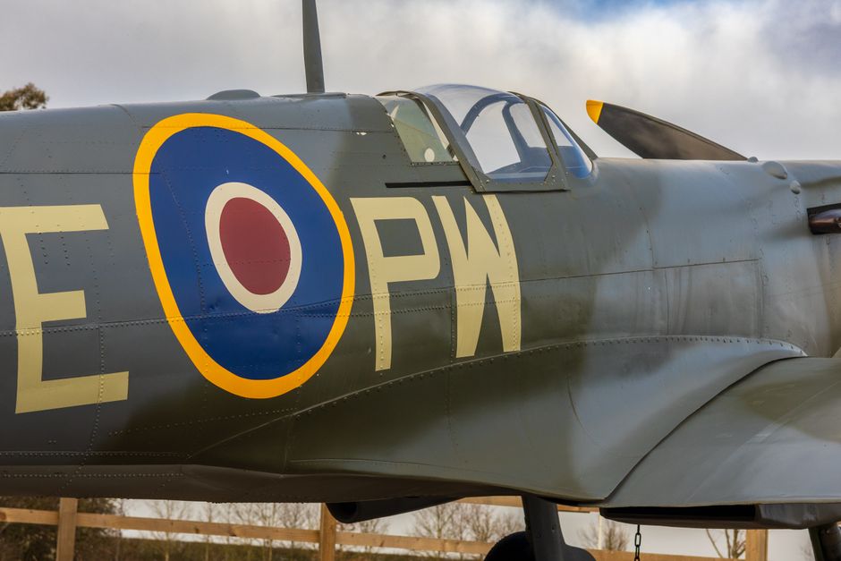 A close-up view of a vintage model aircraft, featuring a metallic green fuselage with visible signs of wear and paint chipping on the surface, revealing a lighter, raw material underneath. The cockpit